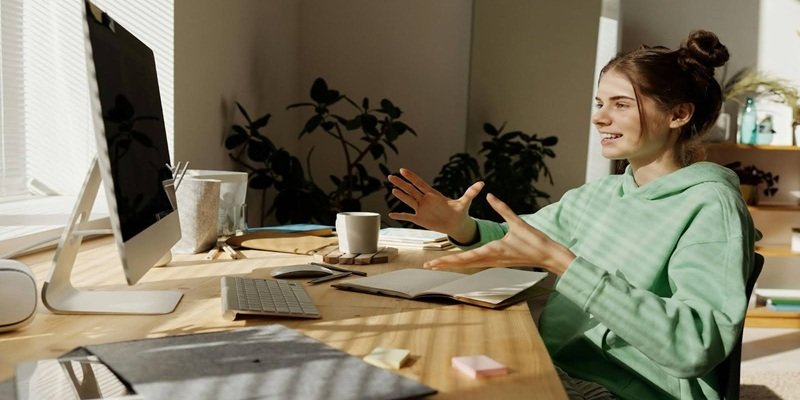 teen girl in Green Hoodie Sitting Near the Table while Having Conversation Through Her Desktop